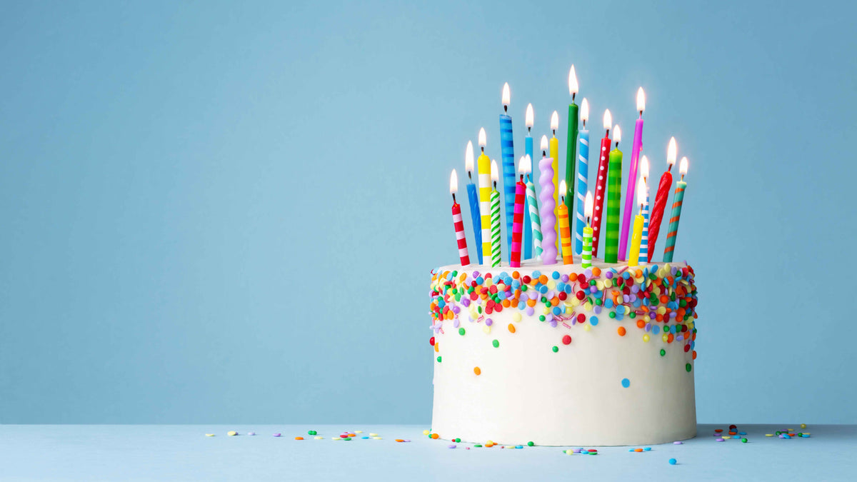 Birthday cake with colourful sprinkles with lots and lots of different colours, patterned, and shaped candles on top. Light pastel blue background.